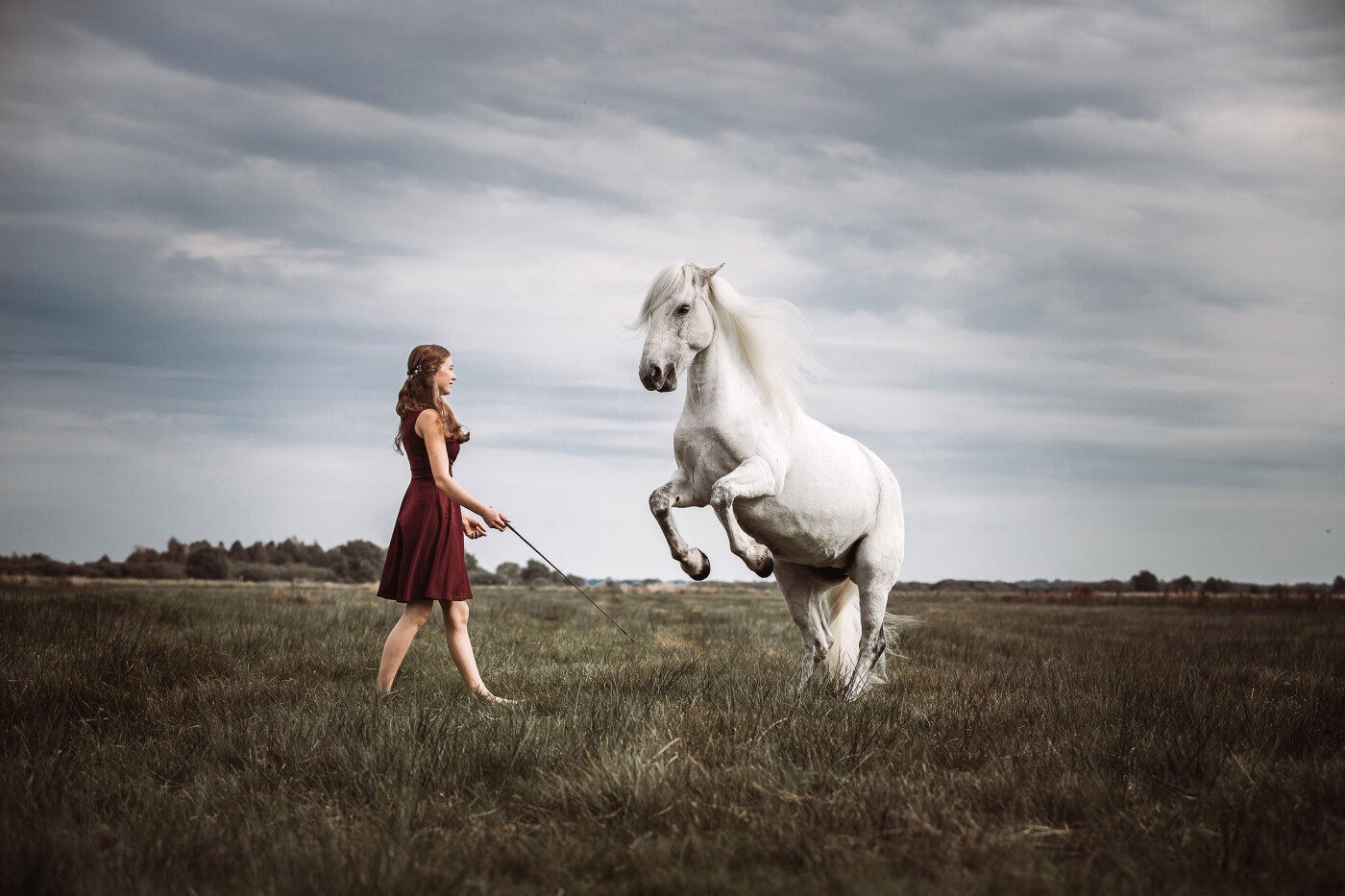 Eine junge Frau in einem Knielangen roten Kleid mit einem vor ihr steigendem weißen Pferd auf einer...
