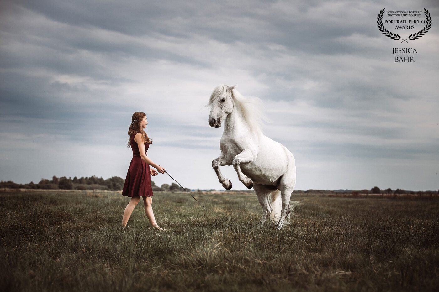 Eine junge Frau in einem Knielangen roten Kleid mit einem vor ihr steigendem weißen Pferd auf einer weiten Landschaft und bewölktem Himmel