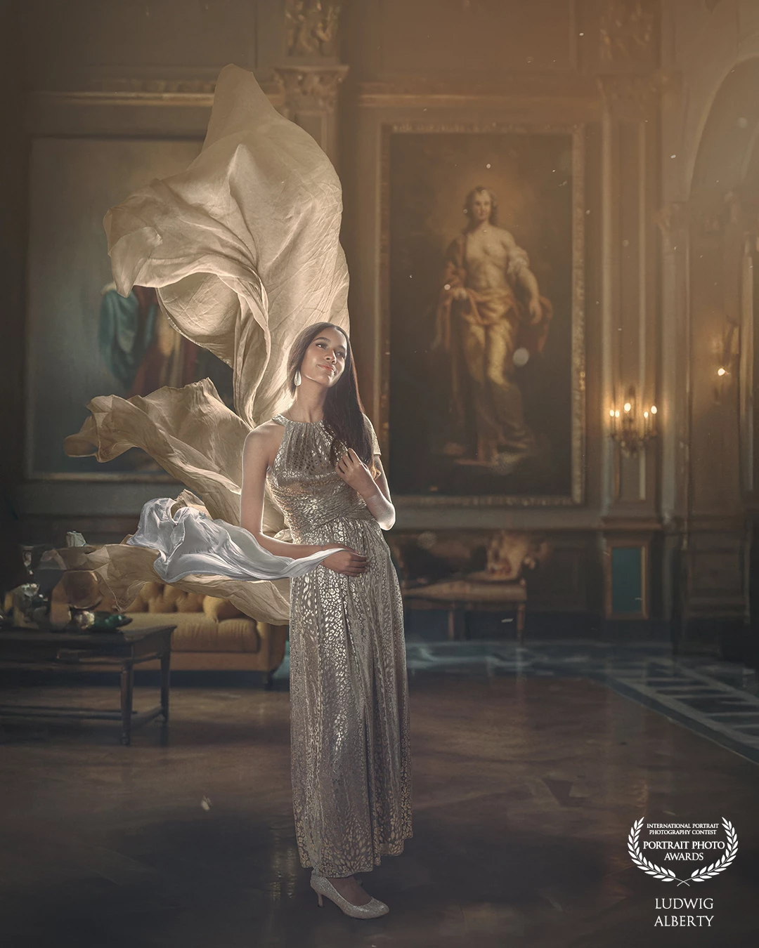 A graceful woman stands in a grand, classical room, wearing a shimmering, sleeveless gown with intricate metallic patterns. She is surrounded by flowing fabric, almost as if caught in a gentle breeze, adding a sense of motion and fantasy. Behind her are large, classical-style paintings and ornate furnishings, suggesting a palace or luxurious historic estate. The lighting is warm and ethereal, casting a soft glow that enhances the fairytale-like atmosphere.