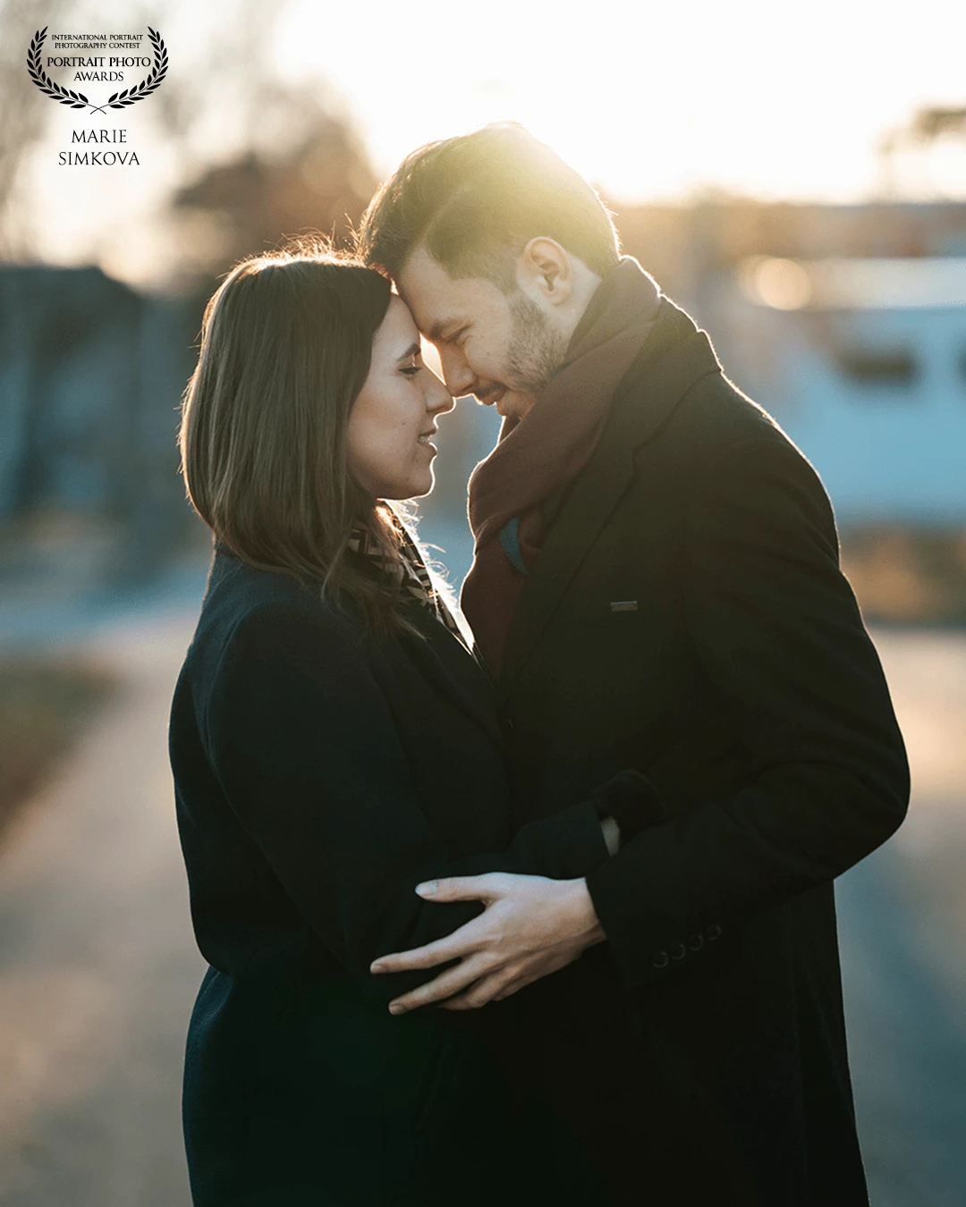 A loving couple stands close, their foreheads touching.<br />
She wears a dark coat and has shoulder-length brown hair; he, in a similar coat and a burgundy scarf, has short dark hair and a beard. Their similarities connecting them even more.<br />
Their hands are gently clasped, with soft sunlight casting a warm glow over them. The blurred background suggests a calm outdoor setting. Making the scene more romantic and in the moment