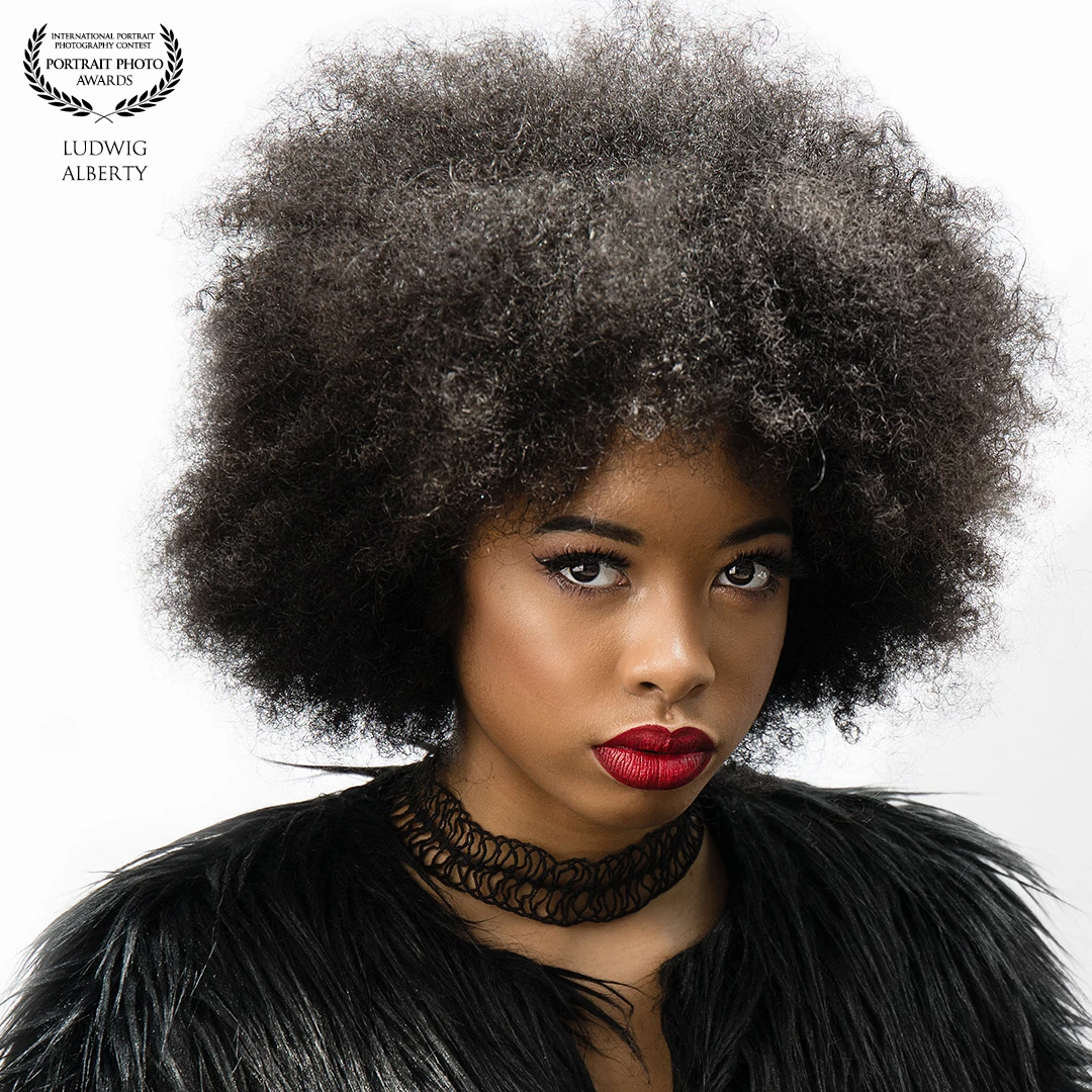 This is a striking studio portrait of a woman with a bold and confident presence. She has a voluminous natural afro hairstyle and is wearing vivid red lipstick, which contrasts beautifully with her smooth, glowing skin. Her makeup is polished, with defined eyebrows, winged eyeliner, and long lashes that enhance her captivating gaze directed at the camera.<br />
<br />
She is dressed in a textured black outfit that resembles faux fur or feathers, adding a dramatic and high-fashion feel to the image. Around her neck, she wears a unique black choker that contributes to the edgy, stylish look. The background is a clean white, which helps to emphasize her features and styling.