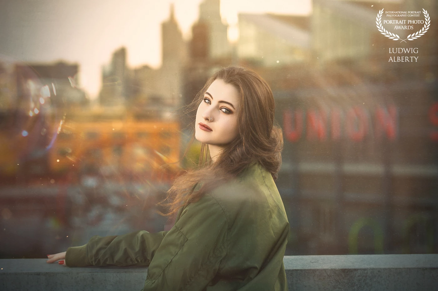 The image depicts a young woman with long, flowing brown hair, leaning against a ledge with a cityscape blurred in the background, suggesting an urban setting during sunset or sunrise with warm orange and pink hues. features a dreamy, ethereal quality with light flares and bokeh effects.