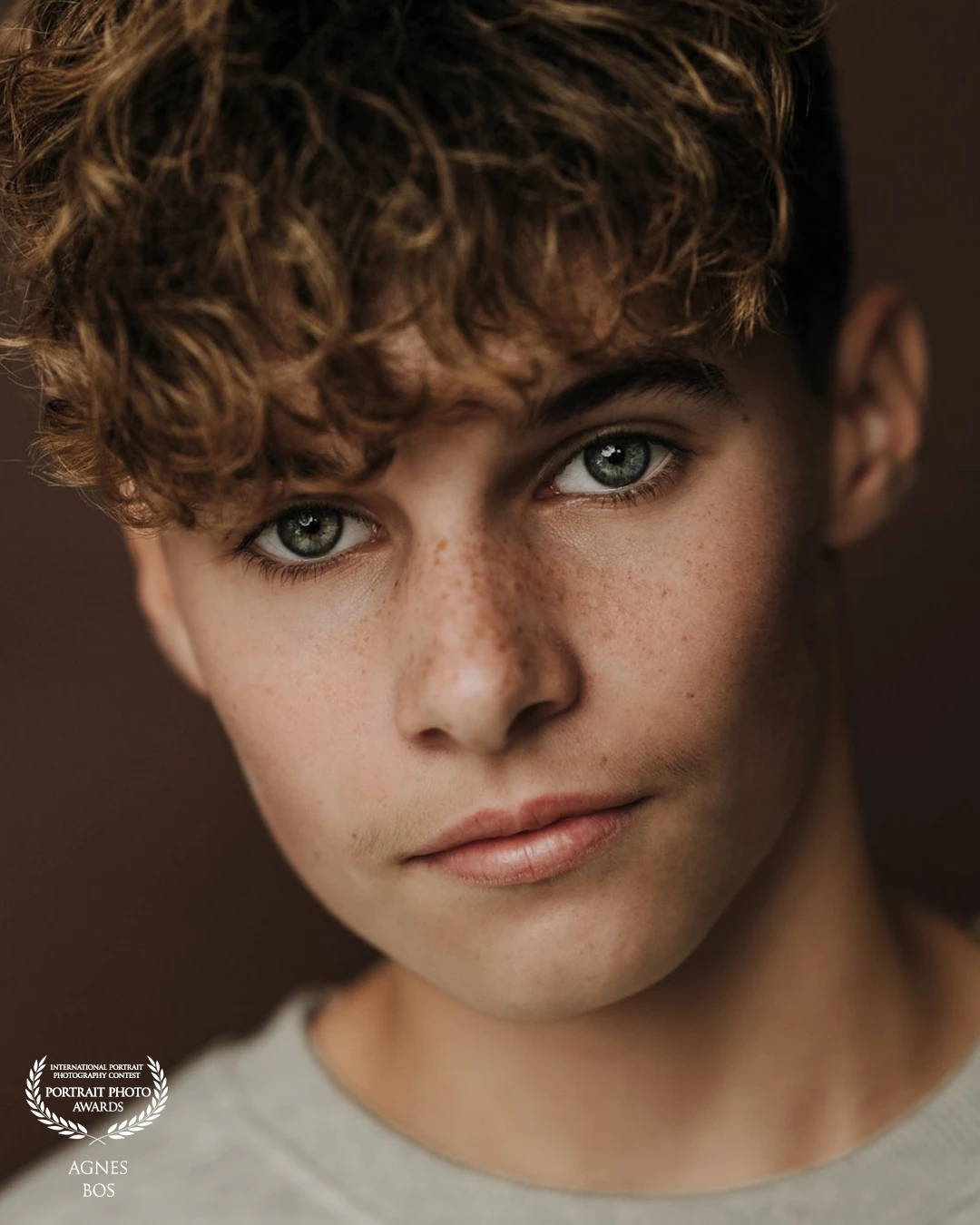 “A timeless pure portrait captured against a warm brown backdrop. His striking curls and piercing blue eyes draw you in, while the simplicity of the composition keeps the focus entirely on his natural beauty and expression.”