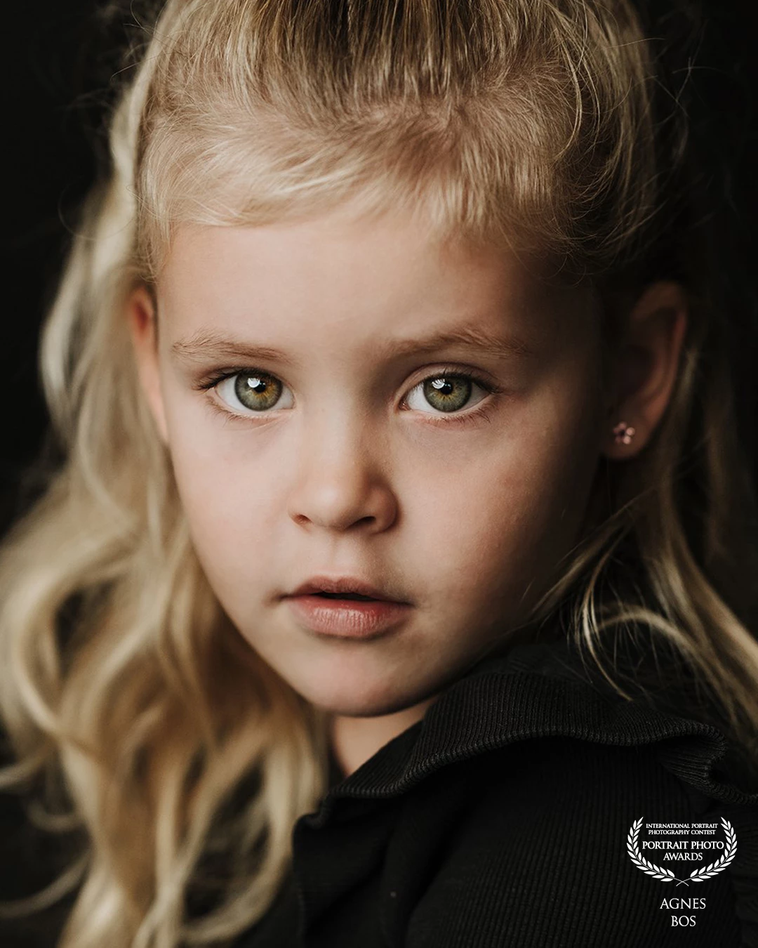 “A pure portrait of a young girl. Her gaze is powerful and unforgettable, set against the timeless contrast of a black background.”