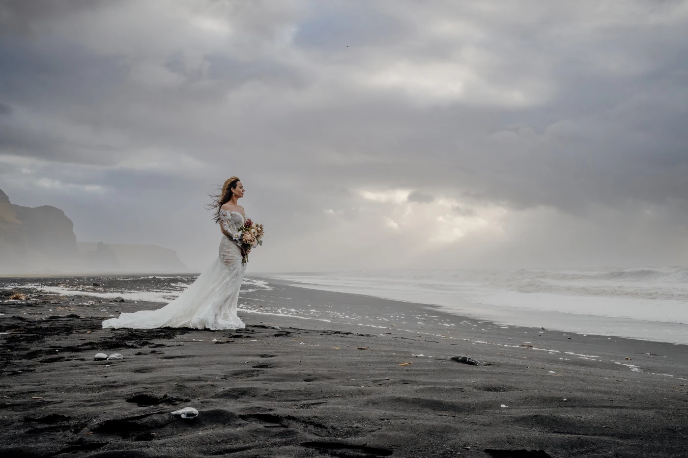 On Iceland’s iconic black-sand shore, she stands quietly against the storm — a lone figure wrapped i...